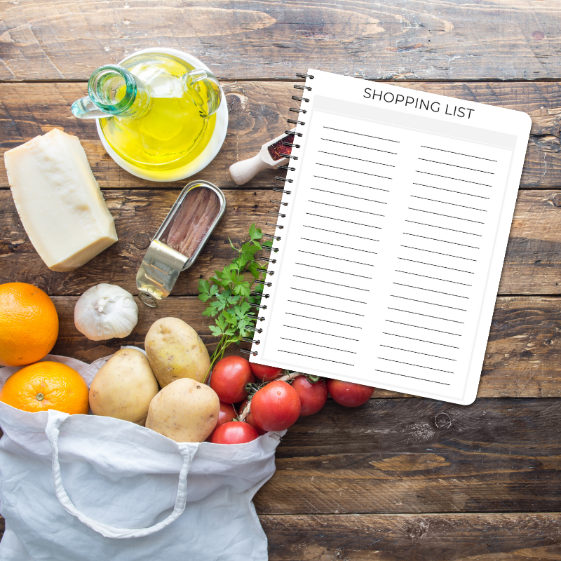 Flat lay of Shopping List page on a wooden background with fresh produce, herbs, olives, and oil.