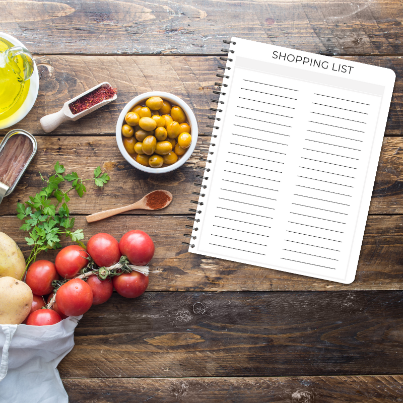 Flat lay of Shopping List page on a wooden background with fresh produce, herbs, olives, and oil.