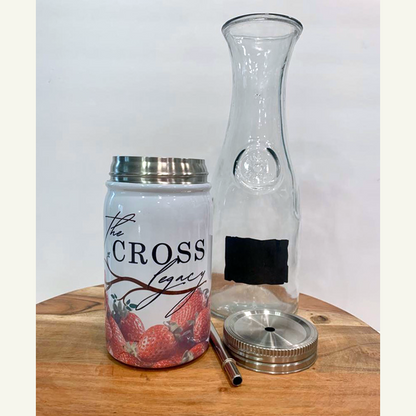 the-cross-legacy-stainless-steel-tumbler-sitting-on-a-wooden-table-with-the-lid-and-straw-laying-on-the-table-and-an-empty-carafe-white-background